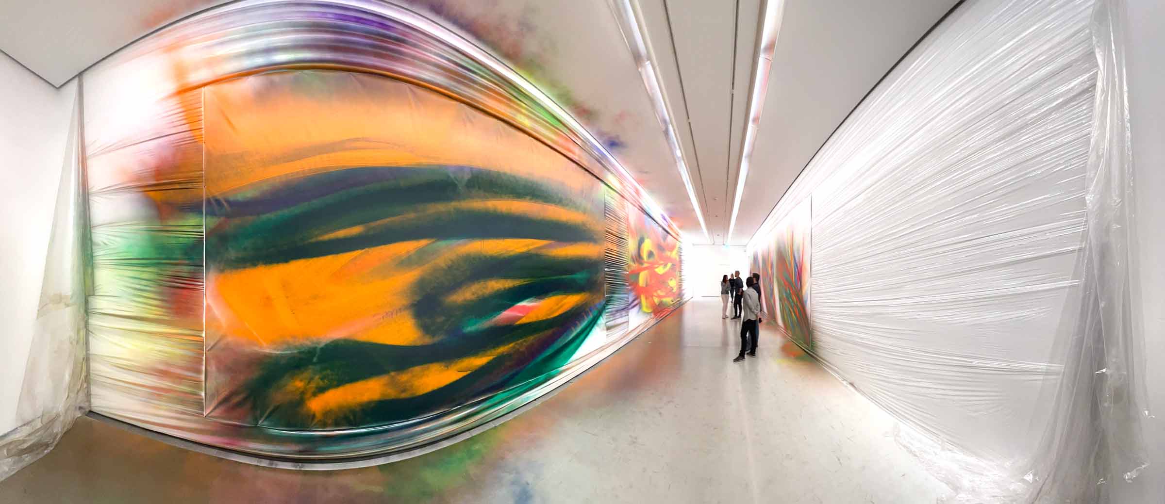 Ausstellungsraum im Weitwinkel mit bunten Kunstwerken und gewellten Folien an der Wand, auf denen sich die Farbe fortsetzt